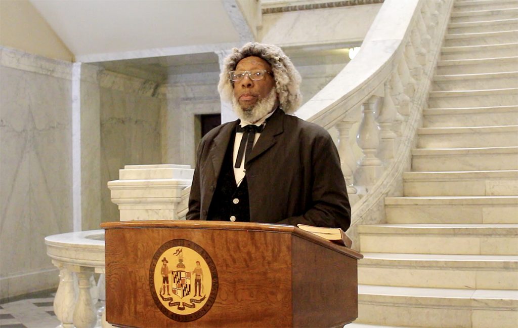 Frederick Douglass interpreter stands at a podium with the logo of the Maryland State House on it. Behind him are marble white stairs. 